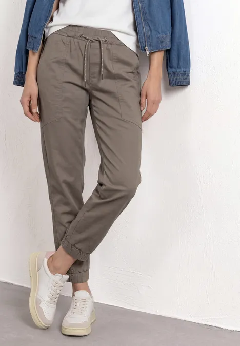 Persoon draagt comfortabele grijsbruine broek met elastische taille en witte sneakers voor een witte muur.