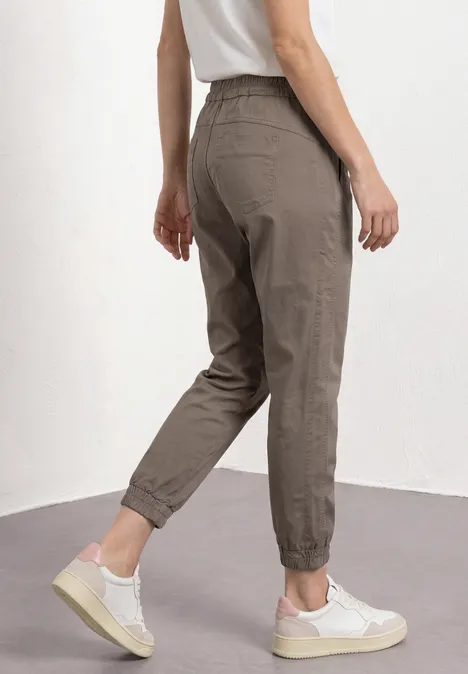 Een vrouw draagt taupekleurige broek met elastische enkels en witte sneakers.
