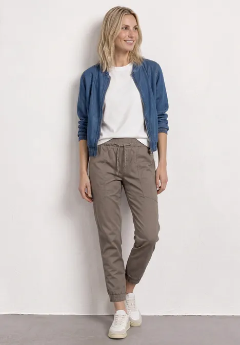 Vrouw in wit t-shirt, blauw jasje, grijze broek en witte sneakers tegen een neutrale achtergrond.