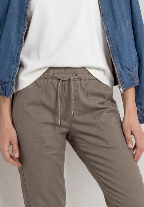 Iemand draagt een taupekleurige broek met trekkoord, een wit t-shirt en een spijkerjasje.