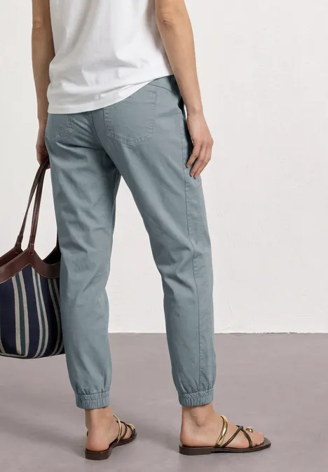 Person von hinten in grauen Hosen mit Gummibund, weißem Oberteil, gestreifter Tasche und flachen Sandalen.