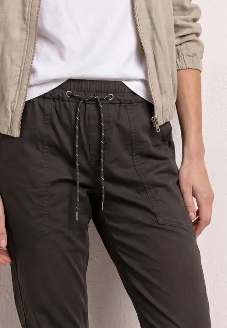 Eine Person trägt schwarze Leinenhose mit Tunnelzug und Eingrifftaschen, kombiniert mit weißem T-Shirt und beigem Hemd.