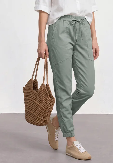 Frau trägt grüne Stoff-Jogginghose mit Bündchen, weißes Hemd, Basttasche und beige Sneaker.
