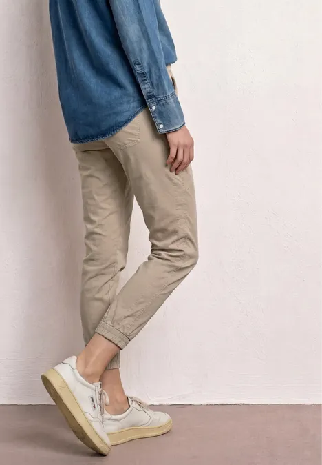 Mann lehnt an Wand, trägt hochgekrempelte beige Chinos, Jeanshemd und weiße Sneaker.