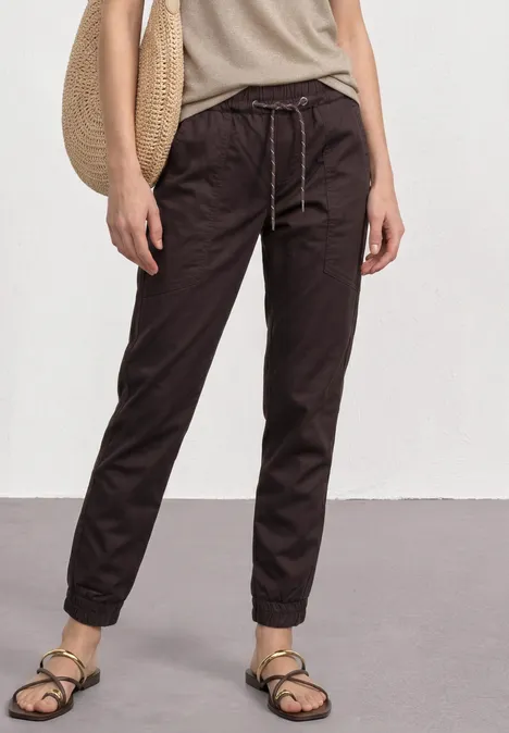 Person in lockeren, dunklen Joggpants mit Kordelzug, beigen Sandalen und Strohtasche.