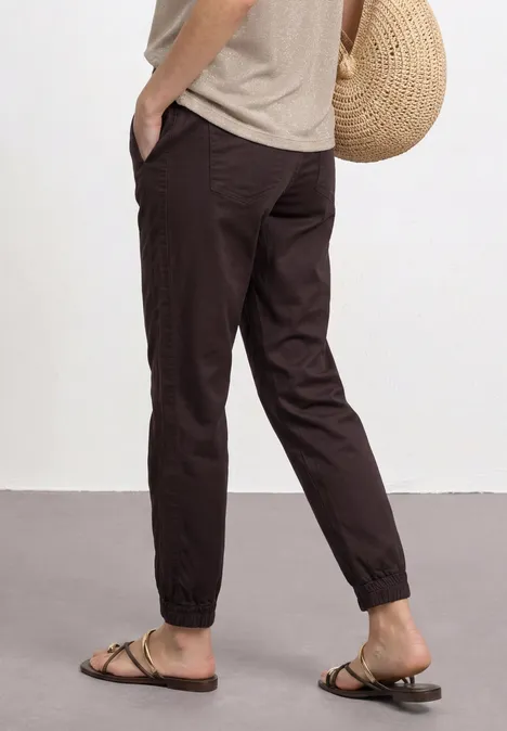 Dunkelbraune, lässige Joggerhose mit Bündchen, kombiniert mit Sandalen, beigem Shirt und Strohtasche.