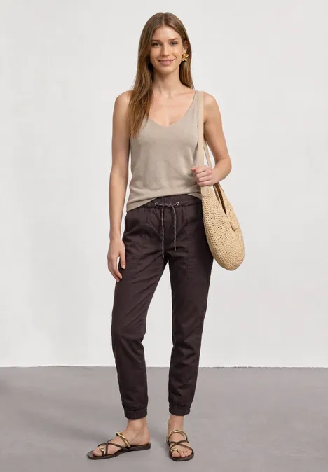 Frau trägt beige Top, dunkle Hose und Sandalen, hält eine Strohtasche.