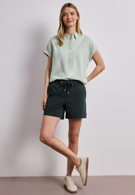 Vrouw in pastelkleurig groen blouse en donkergroene short voor een witte muur.