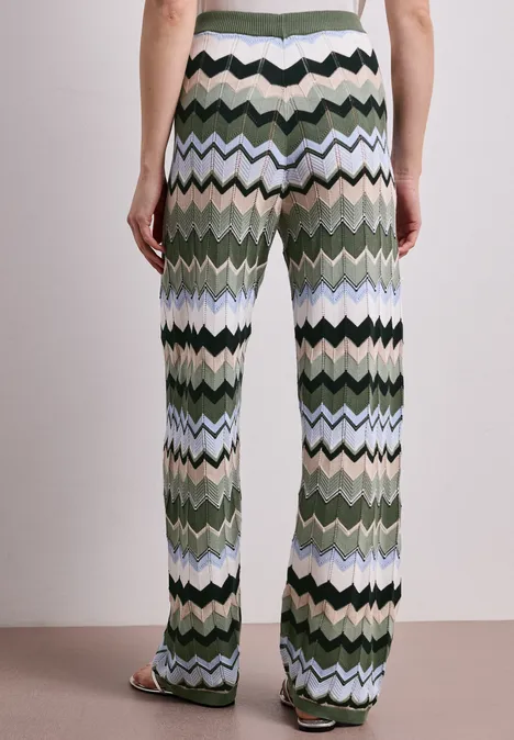 Person von hinten in weiten, gezackten Strickhosen in Grün-, Blau- und Schwarztönen mit offenen Sandalen.