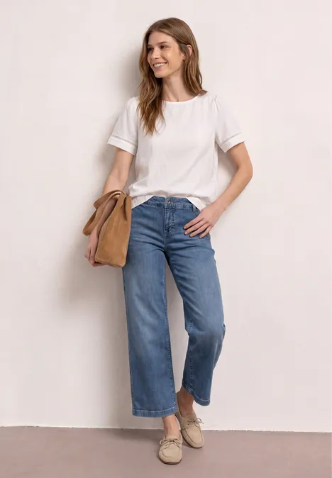 Femme en t-shirt blanc, jean large court, espadrilles et sac besace beige, posant devant un mur clair.