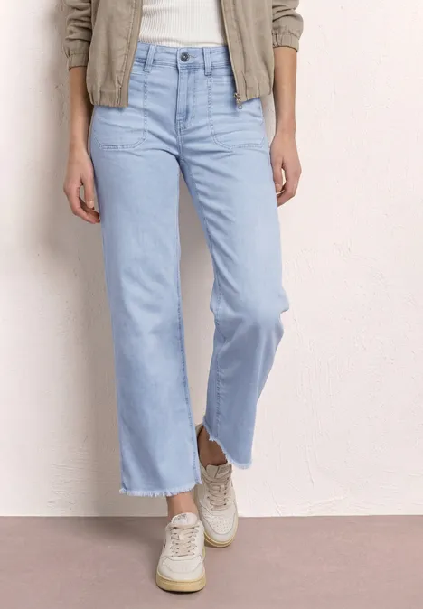 Eine Person trägt helle, ausgestellte Jeans mit fransigem Saum und weißen Sneakern.