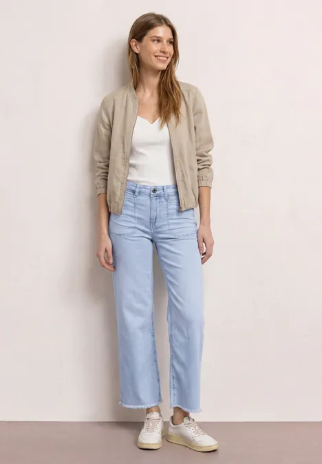 Frau trägt weiße Bluse, beigen Bomberjacke, weite hellblaue Jeans und helle Sneaker.