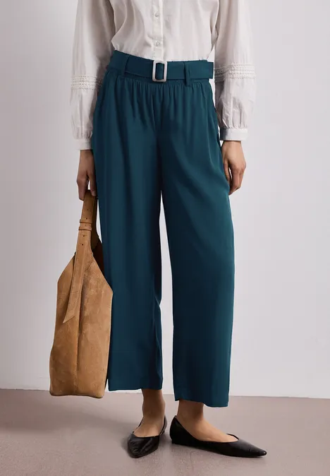 Türkisfarbene, weite Hose mit Gürtel, weiße Bluse, schwarze Ballerinas und beige Tasche.