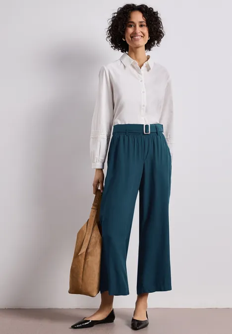 Frau in weißer Bluse, weiten türkisen 7/8-Hosen mit Gürtel, schwarzen Ballerinas und großer Tasche.