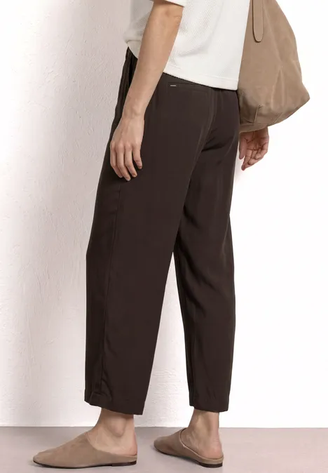 Seitliche Ansicht einer Person mit dunkelbrauner, locker sitzender Hose, beigen Slippern und passender Tasche.