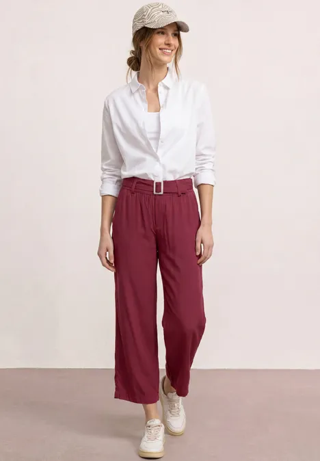 Een vrouw draagt een witte blouse, bordeauxrode broek met riem en witte sneakers voor een lichte achtergrond.