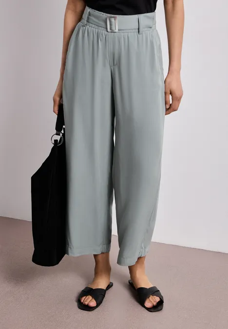 Persoon draagt een wijde, grijze pantalon met riem, een zwart topje, zwarte sandalen en een zwarte tas.