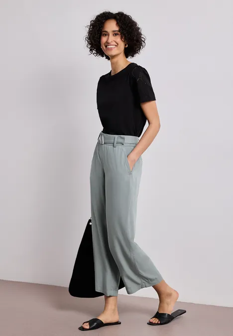 Vrouw in een zwart T-shirt en grijze wijde broek, staand met een zwarte tas en sandalen.