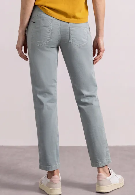 Person von hinten in grauen Jeans, senfgelbem Oberteil und weißen Sneakers.