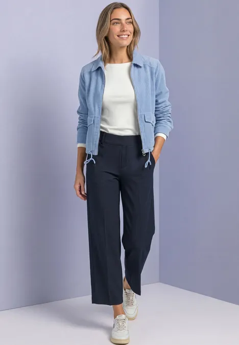 Frau in hellblauer Jacke, weißem Top, dunkelblauen weiten Hosen und Sneakers vor neutralem Hintergrund.