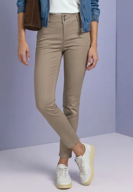 Pantalon chino beige et baskets blanches, avec veste en jean et tee-shirt blanc devant un mur violet.