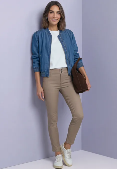 Femme en bomber bleu, t-shirt blanc, pantalon beige et baskets blanches, tenant une pochette marron devant un mur violet.