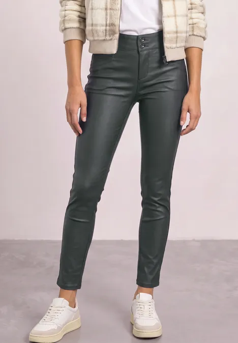Femme en pantalon noir en cuir moulant et baskets blanches, debout devant un fond neutre.