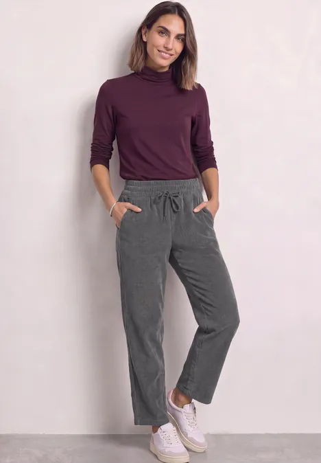 Mid Waist straight leg corduroybroek met rechte pijpen en middelhoge taille gravel grey Een vrouw in een paarse coltrui, grijze joggingsbroek en witte sneakers voor een lichte achtergrond.