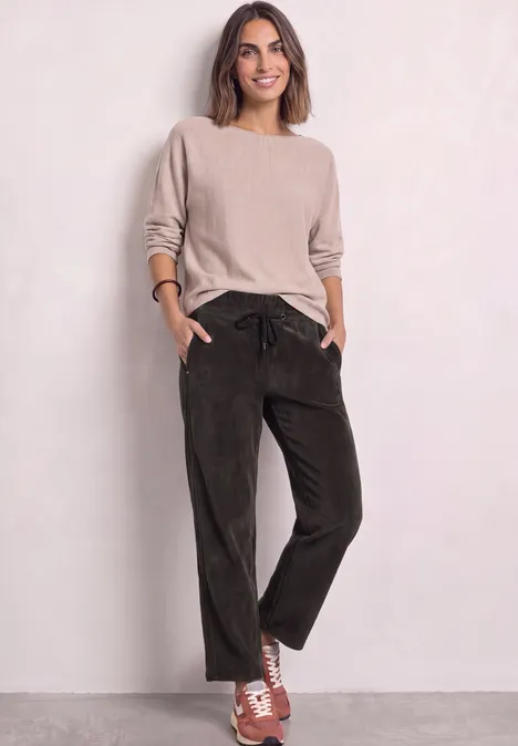 Mid Waist Straight Leg Cordhose in Unifarbe black coffee Frau in beigem Pullover, grünen Cordhosen und orangeweißen Sneakern vor neutralem Hintergrund.