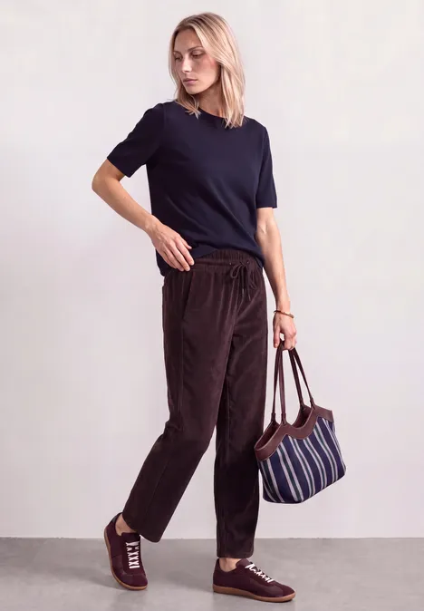 Mid Waist straight leg corduroybroek met rechte pijpen en middelhoge taille vintage ruby Vrouw in donkerblauw T-shirt, bruine corduroybroek en gestreepte tas tegen neutrale achtergrond.