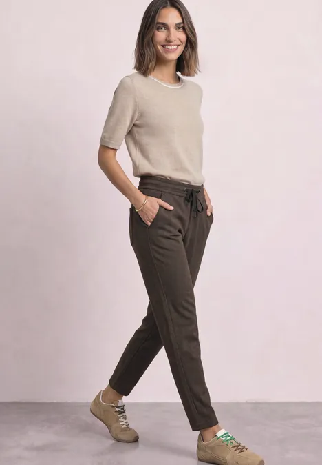 Een glimlachende vrouw in een beige T-shirt, bruine broek en beige schoenen, handen in de zakken.