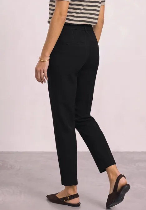 Personne portant un pantalon noir simple et des sandales noires, vue latérale arrière sur fond clair.