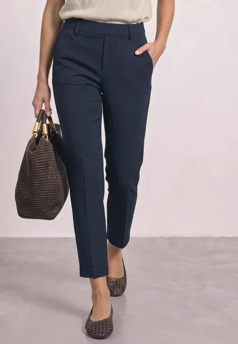 Femme portant un pantalon bleu marine ajusté, des ballerines brunes et tenant un grand sac marron.
