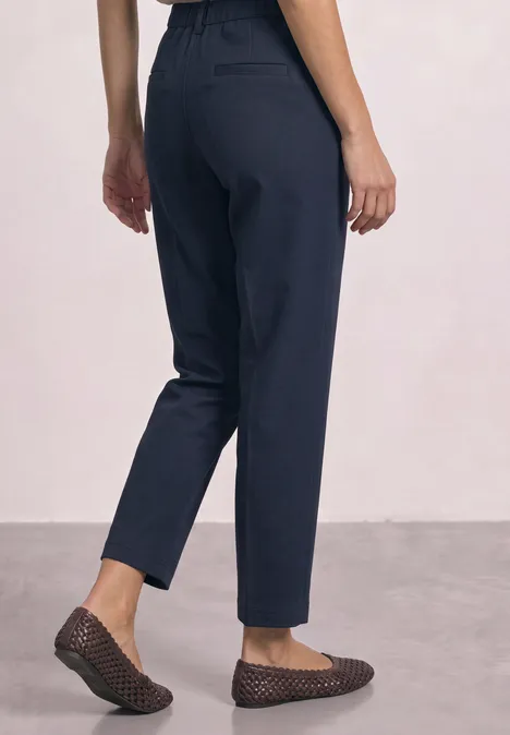 Vue de dos d'une personne portant un pantalon bleu foncé aux chevilles et des mocassins plats sur fond neutre.