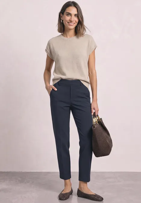 Femme portant un top beige, un pantalon bleu marine et des mocassins, avec un sac à main marron.