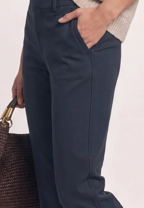 Gros plan d'une personne portant un pantalon bleu foncé et un haut crème, tenant un sac tressé marron.