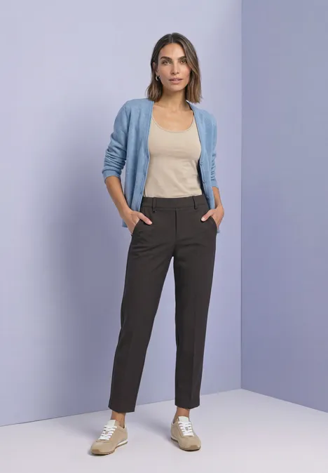 Une femme pose devant un mur violet, portant un gilet bleu clair, un top beige et un pantalon noir.