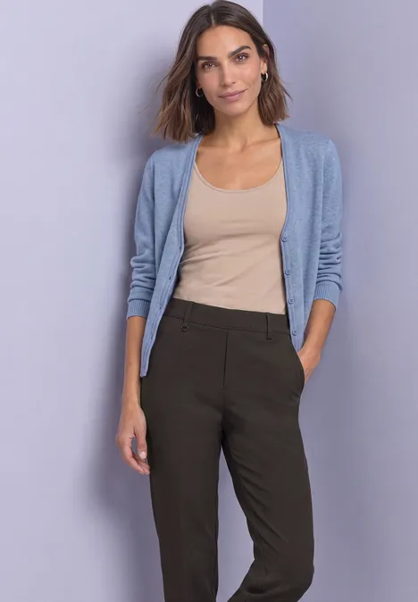 Femme en débardeur beige, gilet bleu clair et pantalon foncé devant un fond uni.