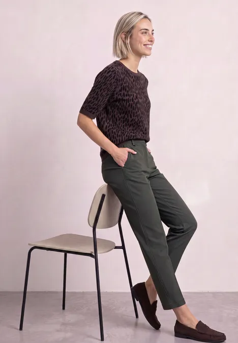 Spodnie chino Slim Leg z brokatowymi akcentami mossbound green Kobieta oparta o krzesło, ma wzorzysty top, zielone spodnie i brązowe mokasyny na różowym tle