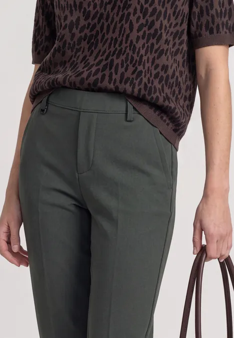 Spodnie chino Slim Leg z brokatowymi akcentami mossbound green Kobieta w zielonych obcisłych spodniach i brązowej, wzorzystej bluzce, trzyma brązową torebkę.