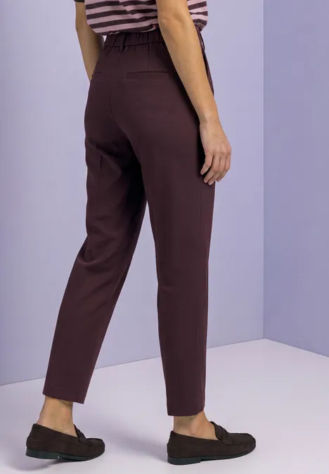 Spodnie chino Slim Leg z brokatowymi akcentami vintage ruby Osoba od tyłu w brązowych, taliowanych spodniach i ciemnych mokasynach na fioletowym tle.