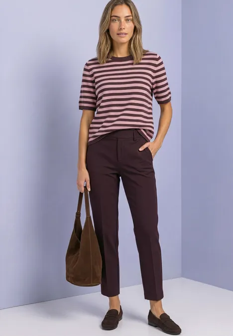 Spodnie chino Slim Leg z brokatowymi akcentami vintage ruby Kobieta w paskowanym t-shircie, ciemnych spodniach, brązowych loafersach i torbie stoi przed fioletową ścianą.