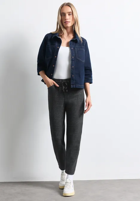 Femme devant un mur blanc, veste en jean, top blanc, pantalon gris et baskets blanches.