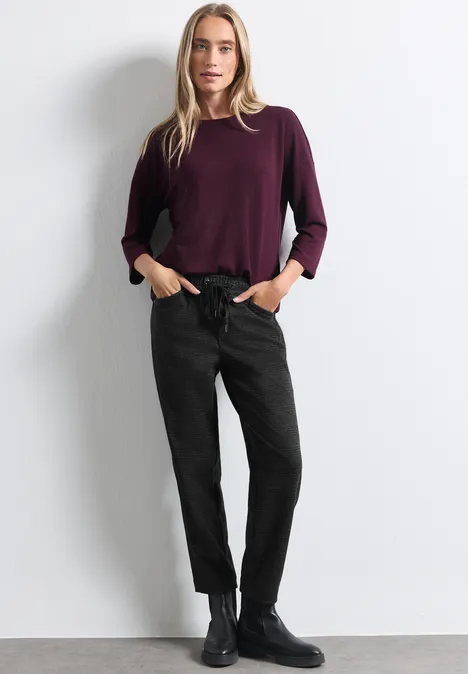 Femme debout sur fond blanc, pull bordeaux, pantalon noir et bottines noires.