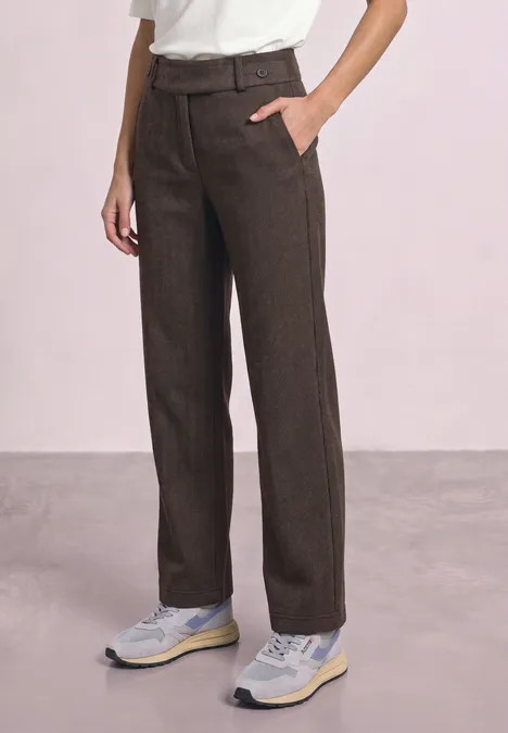 Persoon draagt rechte bruine broek, wit T-shirt en sneakers tegen lichte achtergrond.