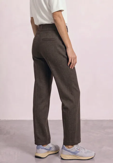 Achteraanzicht: persoon draagt een bruine broek, wit t-shirt en pastelgekleurde sneakers voor een roze muur.