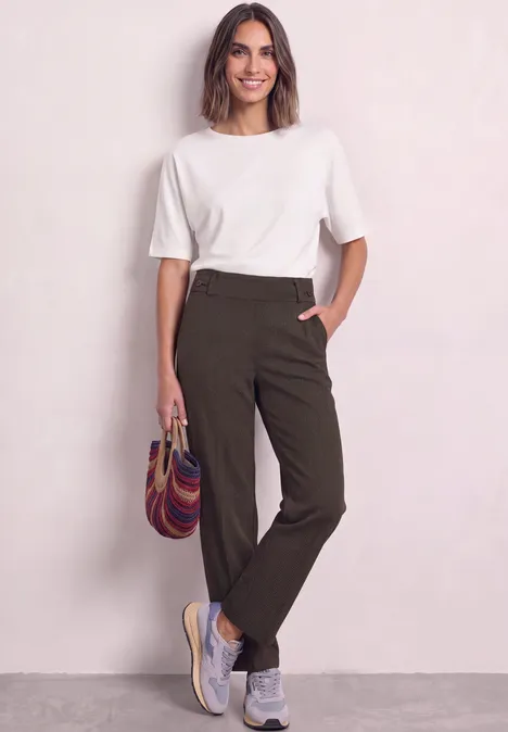 Vrouw in wit T-shirt, groene broek en sneakers, draagt kleurrijke tas voor lichte muur.