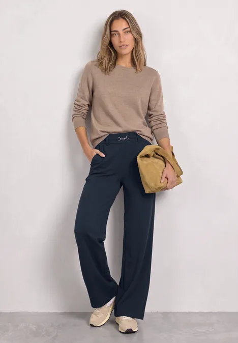 Frau trägt beiges Oberteil, weite dunkelblaue Hose und Sneakers, hält eine beige Tasche.