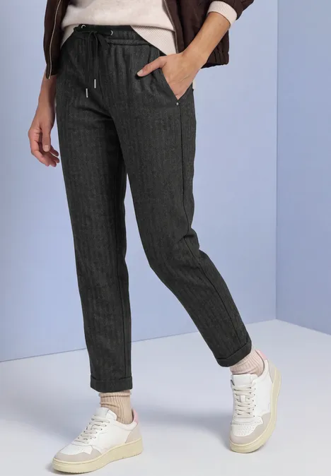 Vrouw draagt donkere joggingbroek met fijne strepen, koordceintuurtje en witte sneakers tegen lichte achtergrond.