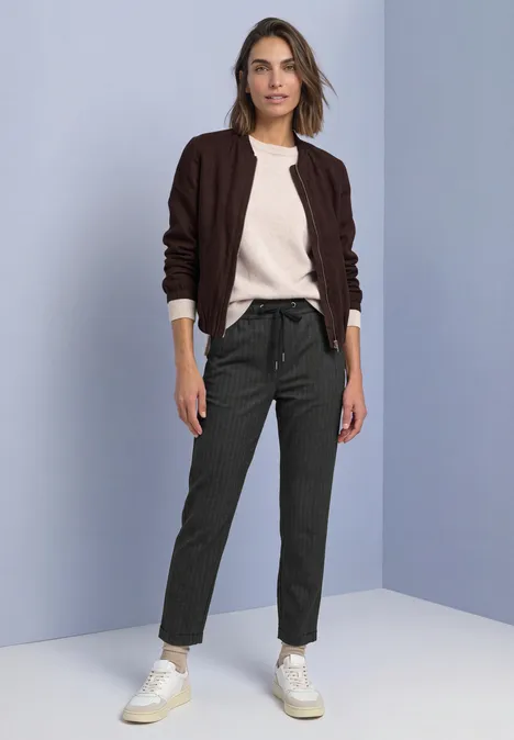 Vrouw in bruine vest, crèmekleurige trui, grijze broek en witte sneakers voor een effen muur.